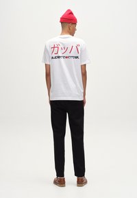 Tricou alb din bumbac cu text roșu în japoneză și „supernormal” în negru pe spate, asortat cu pantaloni negri și pantofi maro.