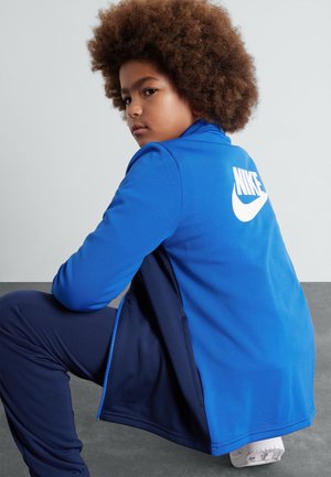 Blå Nike-jacka med vit logotyp, långa ärmar, hög krage och dragkedja. Bärs med matchande blå byxor, vilket visar upp en modern atletisk design.