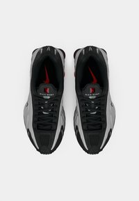 Um par de ténis Nike Shox pretos e cinzentos com realces do logótipo vermelho, vistos de cima sobre um fundo branco.