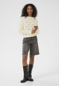 Pull en tricot crème avec un motif en losanges, associé à un short gris foncé et des bottes en caoutchouc noires montantes avec des détails de boucles.