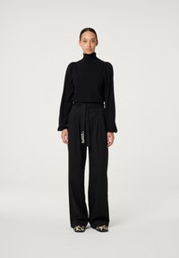 Schwarzer Strick-Turtleneck mit gepufften Ärmeln und schwarzen hoch taillierten Hosen, die mit einem Bindegürtel und dekorativen Akzenten versehen sind. Bedruckte Schuhe runden das Outfit ab.