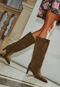 Térd magas, olíva zöld velúr csizmák hegyes orral és stiletto sarokkal, mintázott padló és semleges fal háttér előtt.