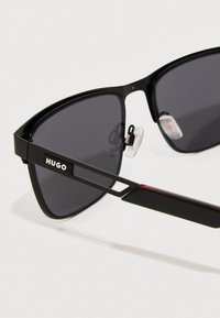 Μαύρα γυαλιά ηλίου με σκούρους φακούς και κομψό ορθογώνιο σκελετό. Διαθέτουν μινιμαλιστική σχεδίαση με το λογότυπο "HUGO" στην πλευρά.