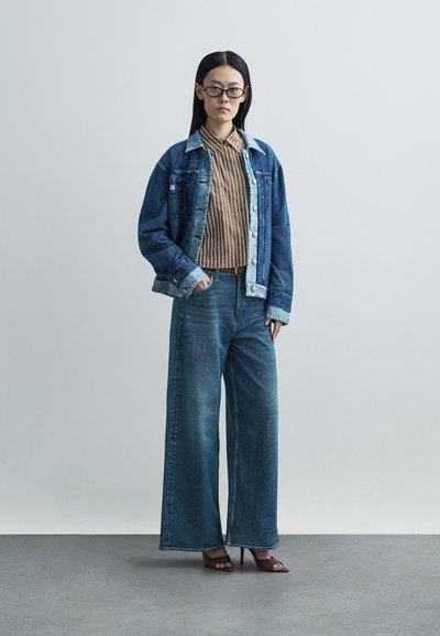 Denimjakk koos kontrastse kraega, triibuline nööbitav särk, laiade säärega denim püksid ja pruunid kontsaga sandaalid, taustal neutraalne taust.