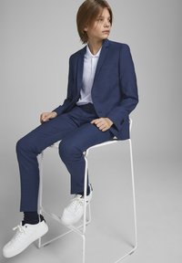 Marineblauw pak met een slim fit, gemaakt van gestructureerde stof, gecombineerd met een wit poloshirt en witte sneakers. Zittend op een witte stoel.