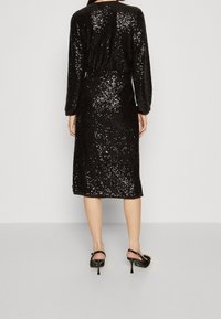 Svart paljetklänning med långa ärmar, mellanhög design, texturerad tyg, samlad midja och diskret glans från paljetterna. Svarta högklackade skor.