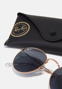 Occhiali da sole Ray-Ban con lenti scure rotonde, montatura in metallo rosa oro e cuscinetti per il naso trasparenti. Include una custodia in pelle nera con dettagli in oro.