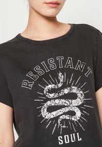 Czarny bawełniany t-shirt z krótkim rękawem, z białym nadrukiem graficznym węża oraz napisem "OPORNY DUCH" na teksturowanej powierzchni.