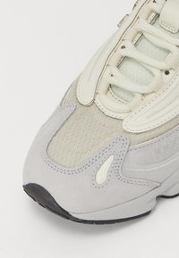 Zapatilla gris y blanco roto con parte superior de malla texturizada, acentos de ante suave y suela de goma con un patrón de tracción distintivo.