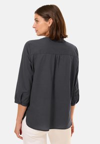 Blusa leggera grigio scuro con maniche a tre quarti, vestibilità ampia, dettaglio arricciato sul retro e polsini bottonati. Tessuto liscio e orlo arrotondato.