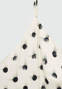 Model cu puncte negre pe o țesătură crem deschis. Dispune de bretele subțiri și o suprafață netedă, ușor texturată.