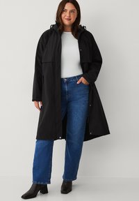 Sort vandtæt frakke med hætte, to forlommer og en løs pasform, båret med blå jeans og sorte hæle støvler. Glat tekstur.