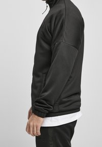 Veste d'athlétisme noire avec col haut, fermeture éclair à l'avant et poches latérales. Texture lisse avec détail de logo sur la manche et la taille.