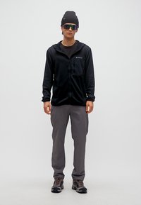 Νεαρός άνδρας με μαύρο σκουφί, ανακλαστικά γυαλιά ηλίου, μαύρο μπουφάν με φερμουάρ, γκρι παντελόνι και παπούτσια πεζοπορίας, στέκεται μπροστά σε απλό φόντο.