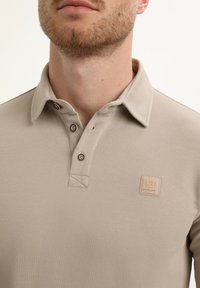 Beżowa koszulka polo z teksturowanej tkaniny, z dwoma brązowymi guzikami i haftowanym kołnierzykiem. Posiada małą metkę z logo na piersi.