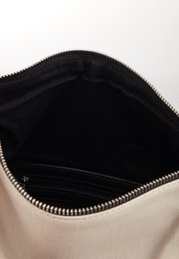 Beżowa torba z zamkiem błyskawicznym, z gładkim tkaninowym wykończeniem na zewnątrz i czarną tkaniną wewnątrz. Zawiera mniejszą kieszeń na zamek błyskawiczny w środku.
