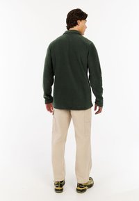 Muškarac stoji okrenut leđima, oblačeći tamnozelenu flis jaknu, bež cargo hlače i žute i crne planinarske cipele, na bijeloj pozadini.