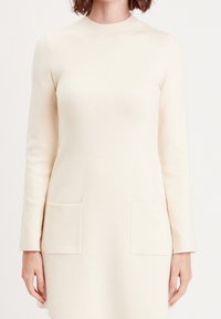 Robe crème à manches longues avec un col montant, dotée de deux poches plaquées à l'avant et d'une texture lisse et douce.