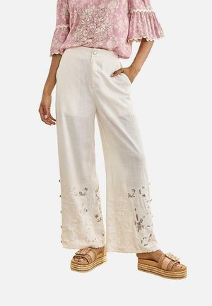 Mujer con pantalones anchos blancos con detalles florales recortados, una blusa rosa bordada y sandalias beige de plataforma.