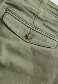 Tissu vert clair en gros plan montrant une poche à rabat boutonnée avec des coutures détaillées sur un vêtement.