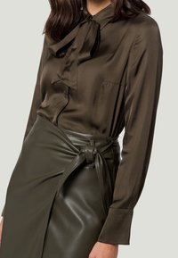 Blouse en soie vert olive foncé avec un nœud au col, associée à une jupe en cuir taille haute avec un détail noué.
