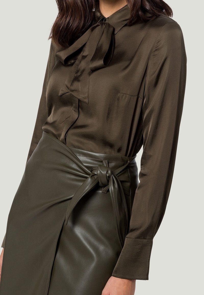 Blouse en soie vert olive foncé avec un nœud au col, associée à une jupe en cuir taille haute avec un détail noué.