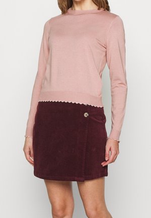 Femme portant un pull à manches longues rose pâle avec des bords festonnés et une mini-jupe en velours côtelé rouge foncé boutonnée.