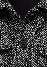 Plašč z vzorcem houndstooth v črno-belem odtenku, izdelan iz teksturirane pletenine, z označenim ovratnikom in zapiranjem na črn gumb.