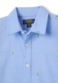 Camicia di cotone azzurro chiaro con colletto, dotata di bottoni bianchi e ricami di palme verdi su tutta la superficie. Etichetta della taglia visibile.