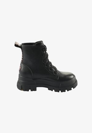 Bota de combate de cuero negro con suela gruesa de tacos, cordones en la parte delantera y una pestaña de tela en el talón con un patrón en rosa y negro.