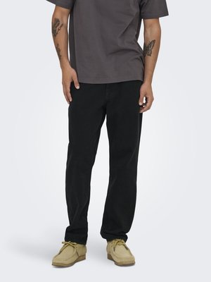 Mann iført svarte bukser, beige mokkasin-sko og en mørkegrå t-skjorte, viser tatoveringer på underarmene mot en nøytral bakgrunn.