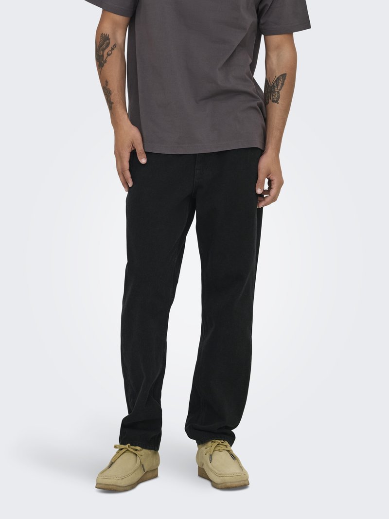 Mand iført sorte bukser, beige mokkasin sko og en mørkegrå t-shirt, der viser tatoveringer på underarmene mod en neutral baggrund.