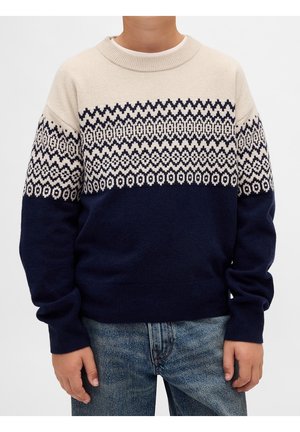 Enfant portant un pull en tricot beige et bleu marine avec des motifs en zigzag et géométriques, associé à un jean bleu, debout sur fond blanc.