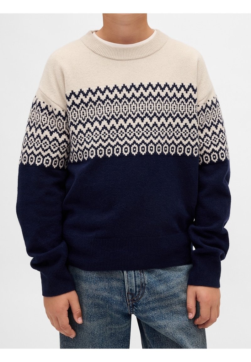 Enfant portant un pull en tricot beige et bleu marine avec des motifs en zigzag et géométriques, associé à un jean bleu, debout sur fond blanc.