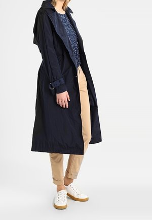 Kvinde iført en navy trenchcoat over en blå mønstret top, beige opsmøgede bukser og hvide sneakers med gummisål.