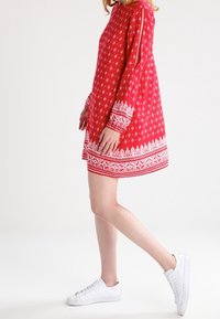 Robe rouge à manches longues, avec motifs floraux blancs et bordure à l'ourlet. Comprend des découpes près des épaules. Associée à des baskets blanches.