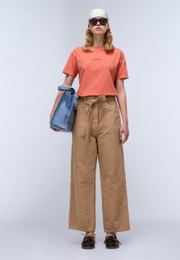 Oranje T-shirt met geborduurde tekst, tan kleurige wijde broek met een gestrikt ceintuur, blauwe handtas en oversized zonnebril, gestyled met een lichte sjaal.