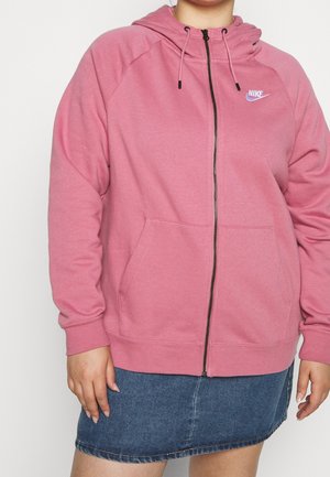Person iført en pink Nike lynlåstrøje med forlommer og et blåt denimskørt mod en ensfarvet baggrund.