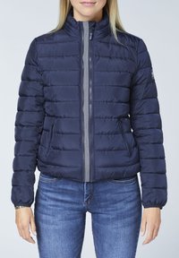 Navy puffer jacket with horizontal quilting, high collar, side pockets, and front zipper featuring striped accents. Model wears blue jeans.