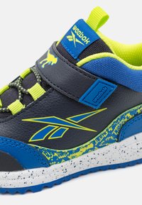 Sneaker nera e blu con accenti gialli, dotata di una tomaia in pelle testurizzata, lacci elastici e una suola bianca maculata. Logo Reebok visibile.