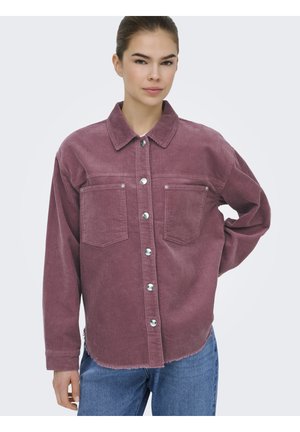 Donna che indossa una camicia di velluto a coste color malva con due tasche sul petto e jeans blu, in piedi con una mano sul fianco contro uno sfondo bianco.