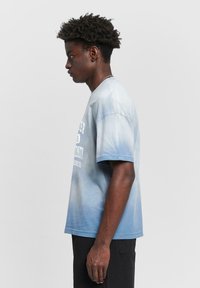 Light blue cotton t-shirt with a gradient fade, featuring short sleeves and a front graphic in white text, worn with black shorts.