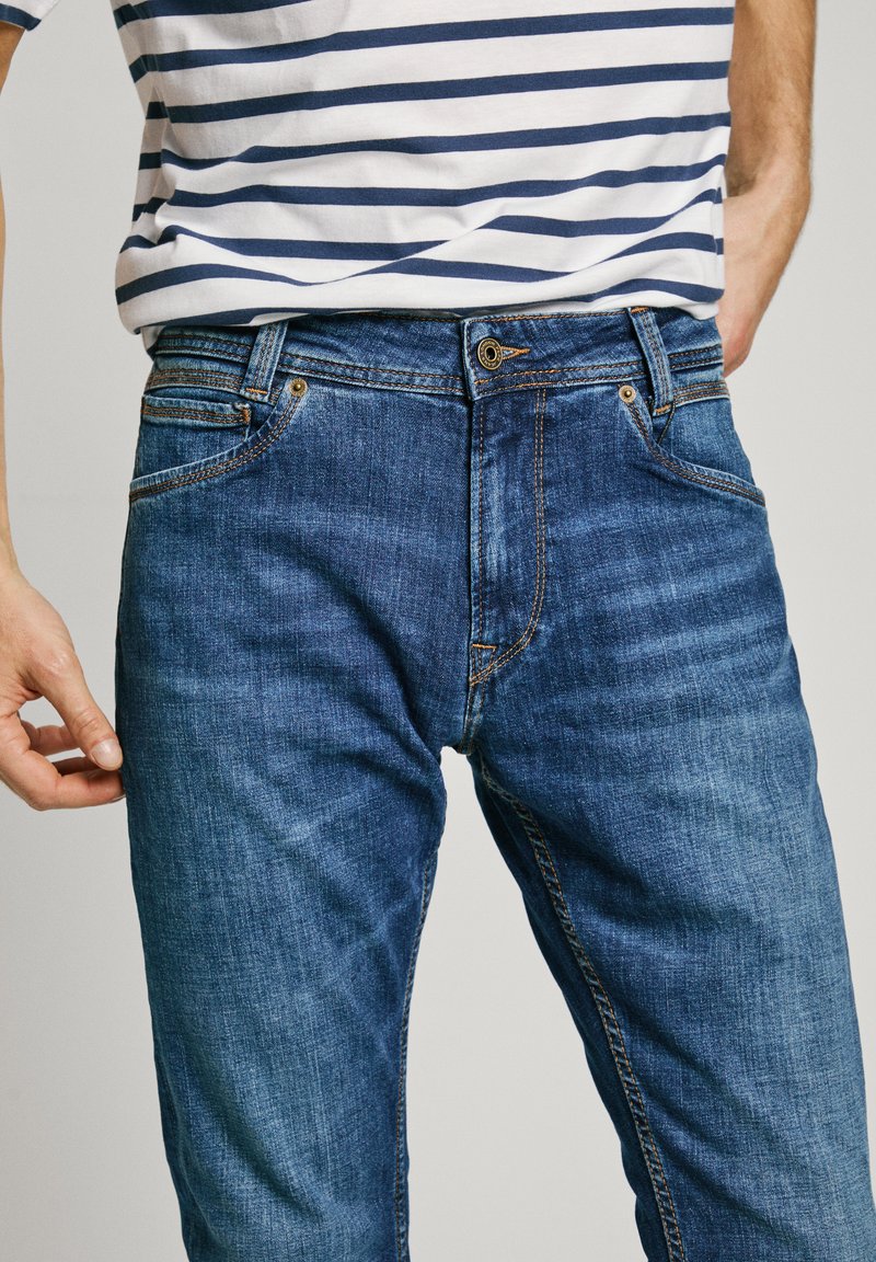Blaue Denim-Jeans mit klassischem Schnitt, ausgestattet mit gestickten Details, fünf Taschen und einem bronzefarbenen Knopf. Weiche Textur und leichtes Ausbleichen.