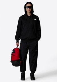 Sweat à capuche noir zippé avec cordons de serrage, associé à un pantalon noir ample. La personne tient un sac à dos rouge et noir, orné d'un logo.