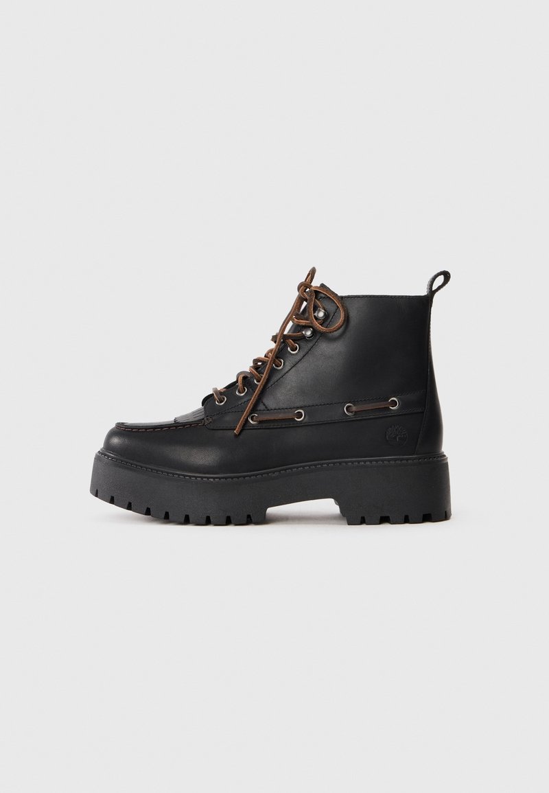 Bottine en cuir noir avec semelle à crampons épaisse, lacets marron, œillets métalliques et languette de traction sur un fond clair uni.