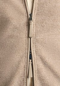 Bež flis jakna z gladkim zadrgo in usnjenim potegom, ki izstopa zaradi mehke teksture in preprostega dizajna.