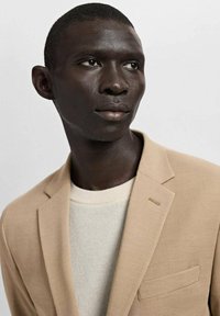 Beiger Blazer mit strukturiertem Design, ausgestattet mit einem Reverskragen und einer einzelnen Brusttasche, kombiniert mit einem hellen, strukturierten Pullover.