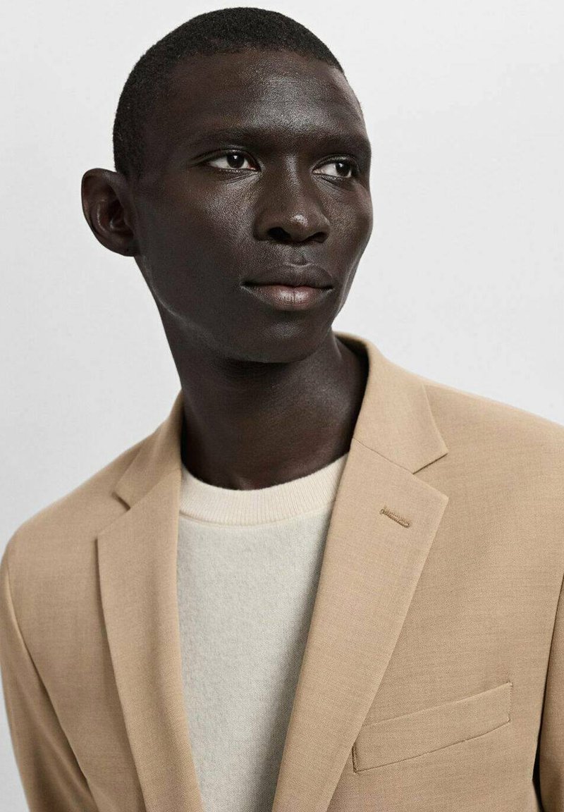 Beige blazer with structured design, featuring a notch lapel and a single chest pocket, layered over a light-colored, textured sweater.