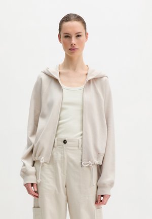 Frau trägt eine hellbeige Kapuzenjacke mit Reißverschluss über einem weißen Tanktop und beige hoch taillierte Hosen, steht vor einem schlichten weißen Hintergrund.