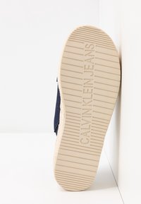 Semelle beige d’une chaussure avec embossage "CALVIN KLEIN JEANS", montrant des lignes de grip horizontales texturées, positionnée contre un mur blanc.